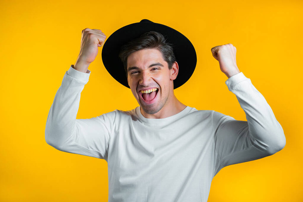 Hipster man in hat is very glad and happy, he shows yes gesture of victory, guy achieved result, goals. Surprised excited happy boy on yellow background - Photo, image