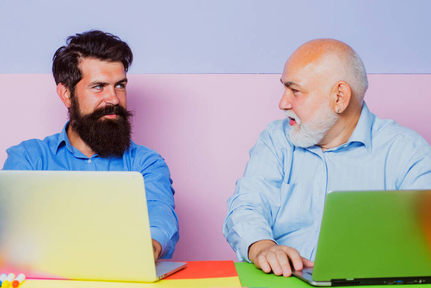 Old and young men looking at laptop screen watching movie or news in social network. Two business people. Business people having meeting around table in modern color office. - Photo, image