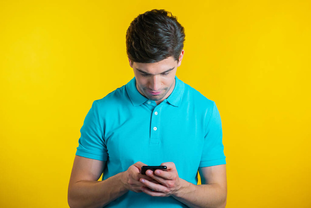 Young man smiling and using mobile phone over yellow background. Handsome european guy holding and texting with smart phone. Boy with technology. - Valokuva, kuva