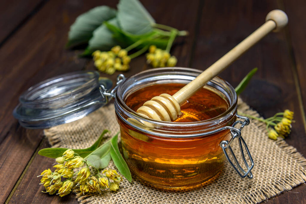 Jar with linden honey with stick for honey and fresh linden flowers on a wooden table. - Photo, Image