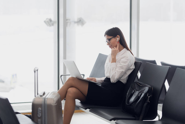 Молодая пассажирка аэропорта на смартфоне и ноутбуке сидит в терминальном зале в ожидании своего рейса. Концепция воздушных путешествий с молодой женщиной, сидящей с ручной клади - Фото, изображение