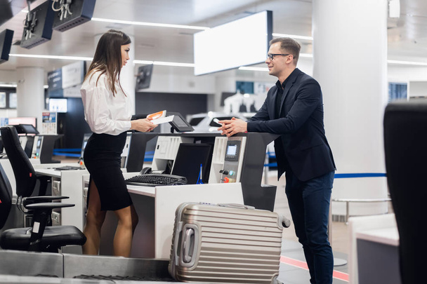 Viagem de negócios. Bonito jovem empresário de terno segurando seu passaporte e conversando com a mulher no check-in da companhia aérea no balcão no aeroporto - Foto, Imagem