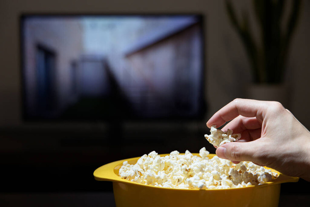 Мужская рука берет попкорн из желтой миски, когда смотрит телевизор - Фото, изображение
