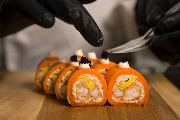Sushi meester serveren Japans Geen rijst roll met tobiko kaviaar op houten snijplank. Chef bereidt voedsel in zwarte beschermende handschoenen. Macro shot van Sushi Roll serveren. Professioneel kookproces - Foto, afbeelding