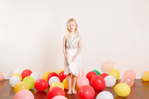 Χαριτωμένο αξιολάτρευτο κορίτσι με λευκό φόρεμα γιορτάζει τα γενέθλια στο σπίτι. Υπέροχο κορίτσι παιδί που διασκεδάζει με πολύχρωμα μπαλόνια. Πάρτι γενεθλίων σε καραντίνα στο σπίτι μόνη της. Κοινωνική απόσταση και αυτο-απομόνωση. - Φωτογραφία, εικόνα