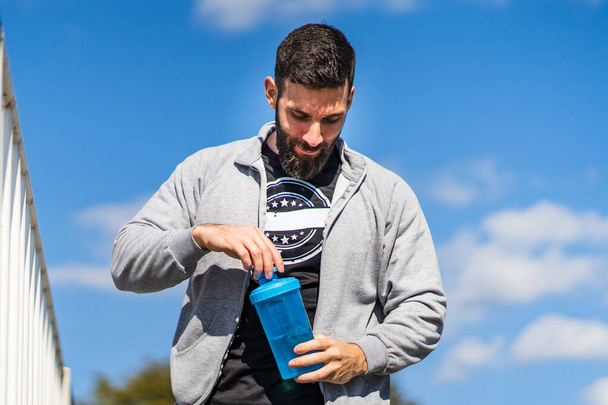 Adult male caucasian athlete standing outdoor with bottle of supplement shaker opening or closing in sunny day - front view waist up copy space - Photo, image