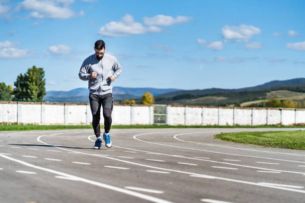 Full length front view on adult caucasian man jogging on the running track - Adult male athlete in stadium training in sunny summer or autumn day - real people healthy lifestyle concept copy space - Photo, Image