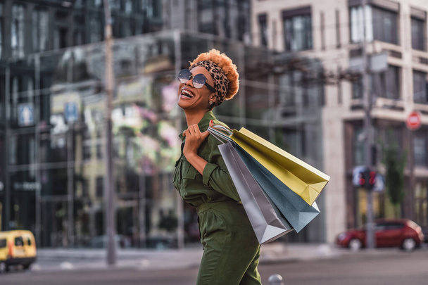 Cheerful attractive Afro American lady walking outdoors while holding shopping bags and laughing. Shopping concept. Copy space - Foto, Bild