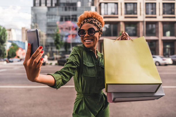 Glücklich modische junge Frau macht Selfie auf dem Smartphone und hält Einkaufstüten in der Hand in der Stadt. Kopierraum. Lifestyle-Konzept - Foto, Bild
