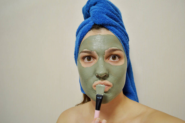Женщина с кистью наносит глиняную маску на лицо с голубым полотенцем на голове - Фото, изображение