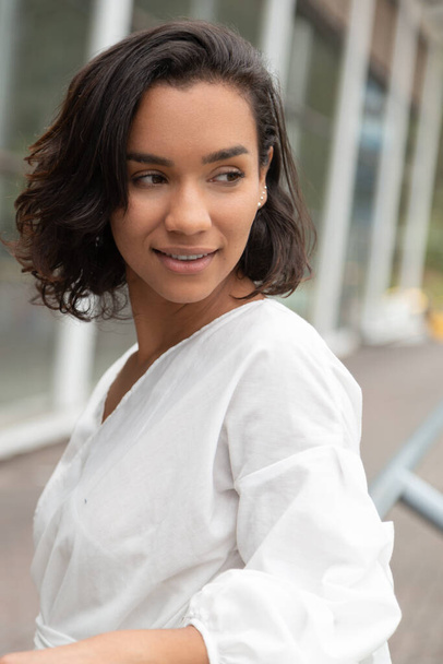 face of young brunette Latin woman with short hair and curls, details of the lips and distracted look, has a white blouse, black pants, outdoors in the day - Photo, Image