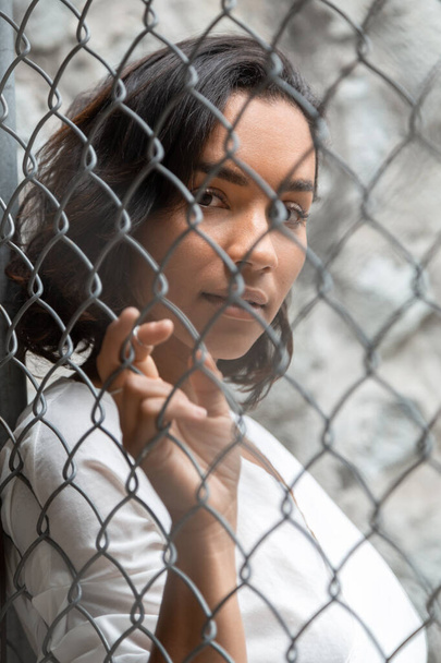 face of young brunette latin girl with short hair and curls, behind a metal mesh, has a white blouse, outside in the day - Fotografie, Obrázek