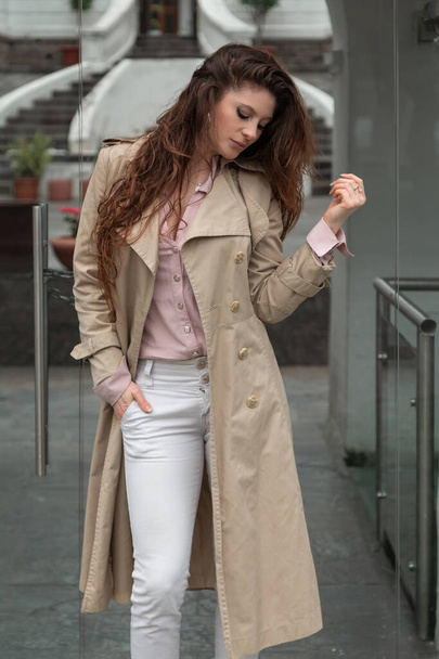 mujer latina joven con el pelo largo y ondulado, de pie en la ciudad, con un abrigo, camisa y pantalones blancos, estilo elegante en el día - Foto, Imagen