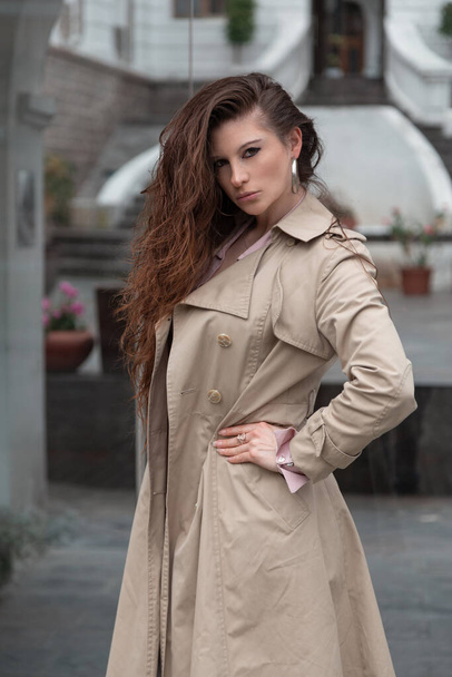 mladá latinská žena s dlouhými vlnitými vlasy, stojící v profilu ve městě, oblečená v béžovém kabátě, ve dne stylová - Fotografie, Obrázek