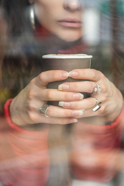 close-up of a person's face holding a cup of coffee with his two hands, in his fingers he has several rings, photo behind glass - Foto, Imagem