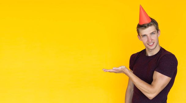young man of European appearance on a yellow background. birthday concept with cap on head, points with hand to blank space for text - Fotoğraf, Görsel