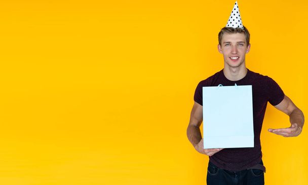 cheerful man on a yellow background with a festive cap on his head. holds a shopping bag in his hands, points at it with his hand - Fotoğraf, Görsel