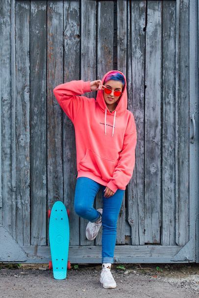 Stylish young woman with short colored hair, dressed in a pink hoodie and pink glasses standing near old wooden gates with her plastic skateboard. Youth concept. - Zdjęcie, obraz