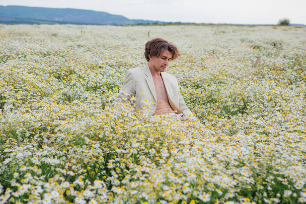 Tall handsome man dressed in a white suit on naked body sitting on a white chair in camomile flowers field - Foto, immagini