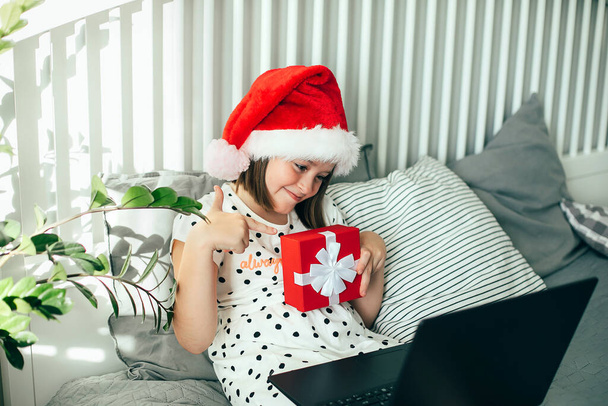 Girl in hat of Santa Claus holding gift box and communicates with friends via video chat. COVID, social distancing concept - Photo, Image