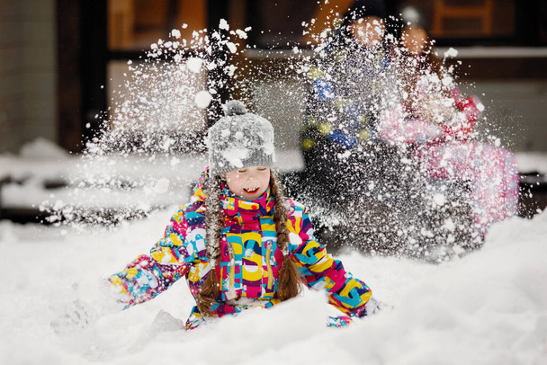 Winter fun, leisure and family vacation concept. Girl with pigtails throws snow in a snowdrift in front of a window at home and family. Selective focus, blurred background. - Photo, Image