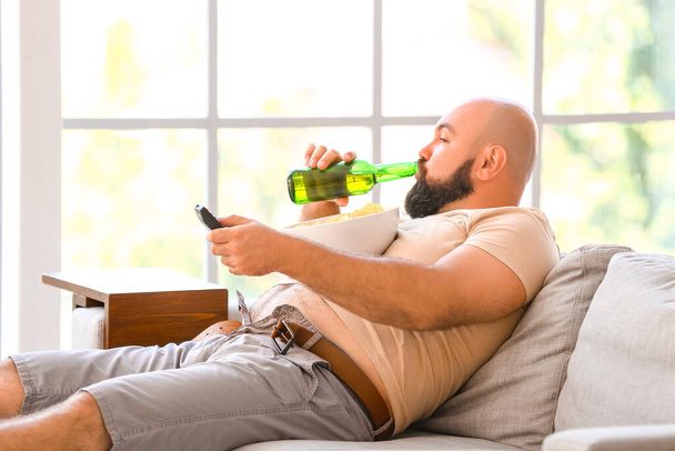 Lazy overweight man with beer and chips watching TV at home - 写真・画像