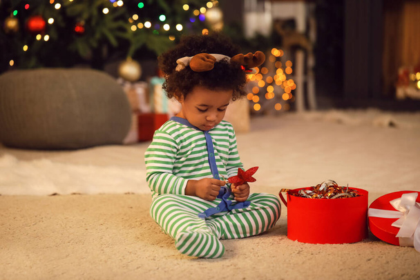 Cute African-American baby girl with gift at home on Christmas eve - Photo, Image