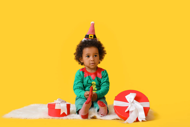 Cute African-American baby girl dressed as elf and with Christmas gifts on color background - Photo, Image