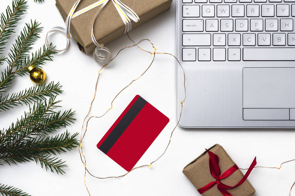 Christmas online shopping. An open laptop on a white background,a credit card, gift boxes tied with a red ribbon, green fir branches, garlands of stars. Online trading during the winter holidays. - 写真・画像