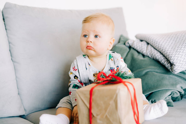 lapsen ensimmäinen joulu. söpö pieni poika istuu juhlapuvussa harmaalla sohvalla, jolla on suuri lahja punaisella nauhalla, valkoista seinää vasten, jossa on luonnollinen kuusen seppele.. - Valokuva, kuva