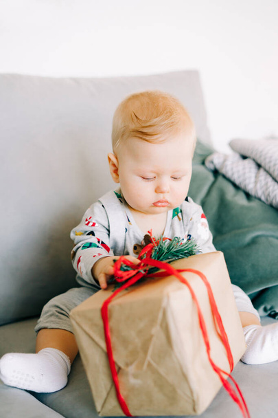 the child's first Christmas. a cute little boy is sitting in a festive costume on a gray sofa with a large gift with a red ribbon, against a white wall with a natural fir wreath.Christmas concept. - 写真・画像