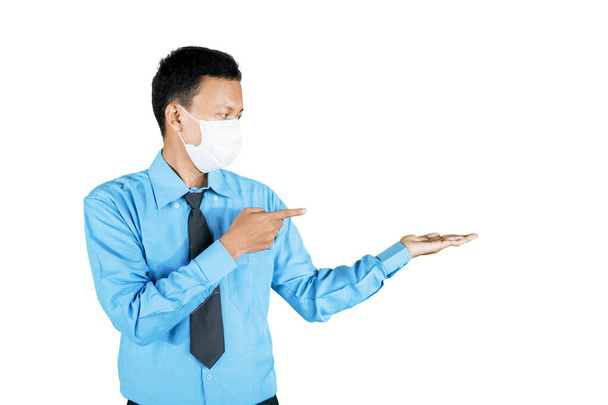 Νεαρός επιχειρηματίας με μάσκα προσώπου ενώ δείχνει κάτι στα χέρια του και στέκεται στο στούντιο. Απομονωμένα σε λευκό φόντο - Φωτογραφία, εικόνα