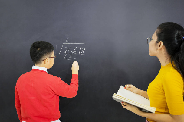 Picture of male student learns math with his female teacher while writing on chalkboard and standing in the classroom - Foto, Imagem