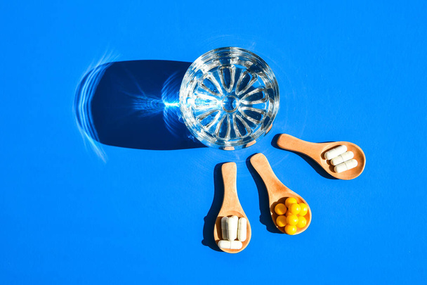 Glas water en pillen op houten lepel op blauwe achtergrond met hard licht en schaduwen. Creatief. Vitaminen en prebiotica, probiotica. Herfst vitamine dosis. - Foto, afbeelding