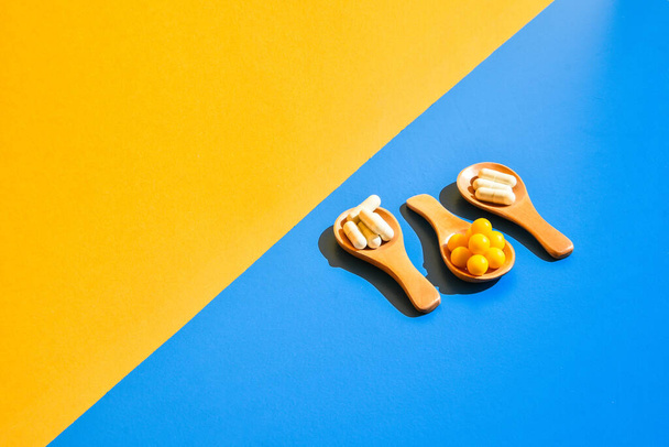 Pillole su cucchiaio di legno su sfondo blu e giallo. Luce e ombre dure. Moderno concetto di minimalismo creativo isometrico. Vitamine e prebiotici, probiotici. Dose di vitamina autunnale. - Foto, immagini