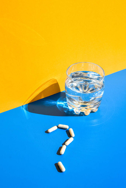 Glass of water and pills in shape of question mark on blue and yellow background. Hard light and shadows. Modern isometric creative minimalism concept. Vitamins and prebiotics, probiotics. Autumn vitamin dose. - Foto, Imagem
