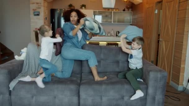 Mixed race lady nanny playing pillow fight with children laughing having fun indoors - Filmagem, Vídeo