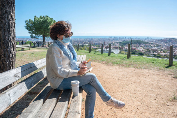 A woman a surgical mask, sitting on a bench writing in her notebook with the city in the background - Fotó, kép