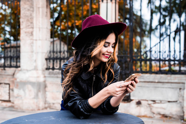 Śmiejąca się dziewczyna ze smartfonem dobrze się bawi w jesienny weekend. Outdoor portret sympatycznej modnej pani z brązowymi włosami nosi kapelusz w październiku dzień. - Zdjęcie, obraz