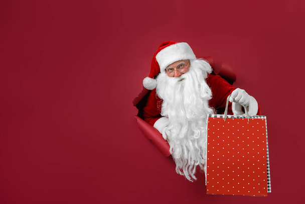 Санта - людина тримає пакети в руці через паперову діру. Одягнений чоловік у капелюсі в Санті дивиться через отвір на червоному папері - Фото, зображення