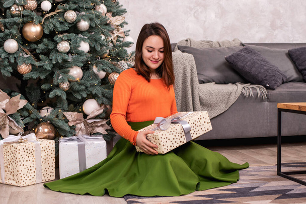 Красивая девушка в оранжевой куртке открывает новогодний подарок в коробке с лентой. Рождество, канун Нового года - Фото, изображение