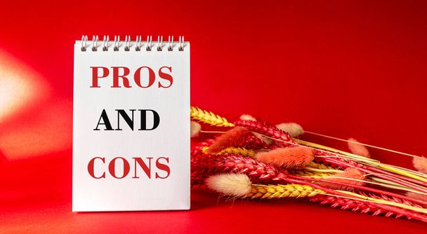Pros and cons, the text is written on a white notebook on a red background framed by spikelets of wheat - Photo, Image