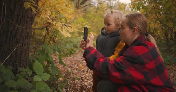 madre está enseñando a su pequeño hijo a tomar una foto por teléfono inteligente una naturaleza, viaje familiar en el bosque o parque de otoño - Imágenes, Vídeo