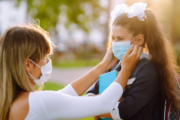 Caring Mother helps to wear a medical mask schoolgirl outdoors. School during the COVID-19 quarantine. - Фото, изображение