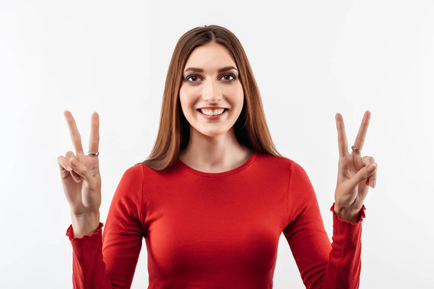 Porträt einer glücklichen jungen Frau mit langen kastanienbraunen Haaren, die eine Friedensgeste zeigt und einen lässigen roten Pullover trägt. Studioaufnahme, weißer Hintergrund. Menschliche Emotionen, Gesichtsausdruck - Foto, Bild
