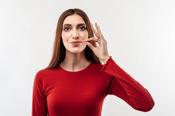 Young woman with long chestnut hair in casual red sweater, showing zip gesture as if shutting mouth on key, promises to keep secret, holds fingers near cheek. Silence and secret concept - Photo, Image