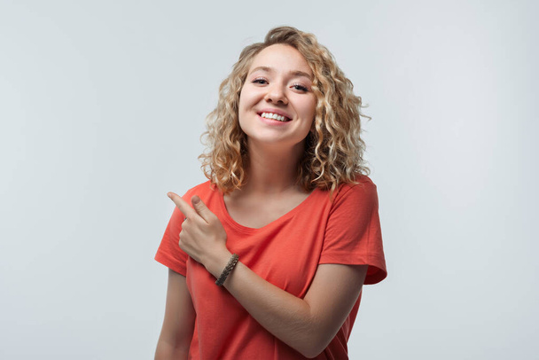 Imagen de una joven mujer feliz con el pelo rizado, vistiendo una camiseta casual, sonriendo y señalando con el dedo índice a un lado, muestra el lugar para el anuncio. Captura de estudio, fondo blanco, aislado - Foto, Imagen