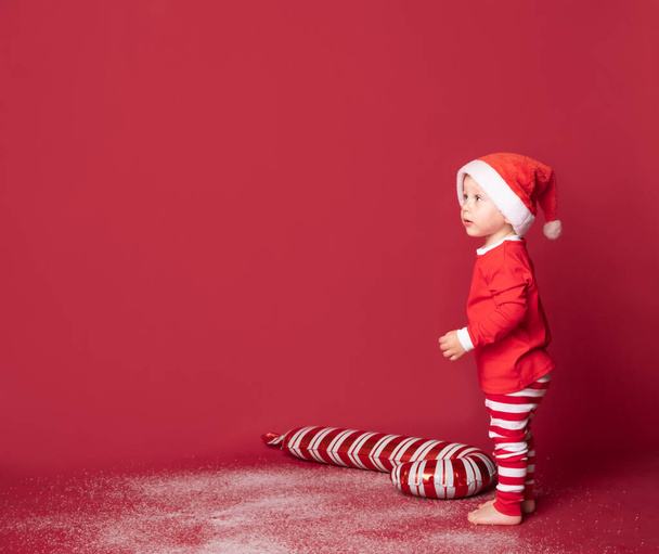 Νήπιο κορίτσι σε Σάντα καπέλο με τα Χριστούγεννα ζαχαροκάλαμο καραμέλα σε κόκκινο φόντο. - Φωτογραφία, εικόνα