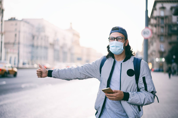 Cute hipster brunette in glasses and a protective mask on his face hitchhiking, in the big city on the background of the road, covid pandemic new normal lifestyle - Photo, Image
