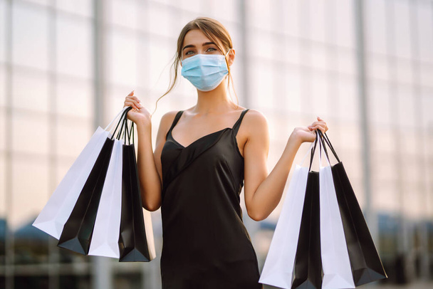Stylish woman in protective medical mask holding shopping bags near mall. Shopping in the coronavirus epidemic. Sale, consumerism, Black friday concept.  - Фото, зображення